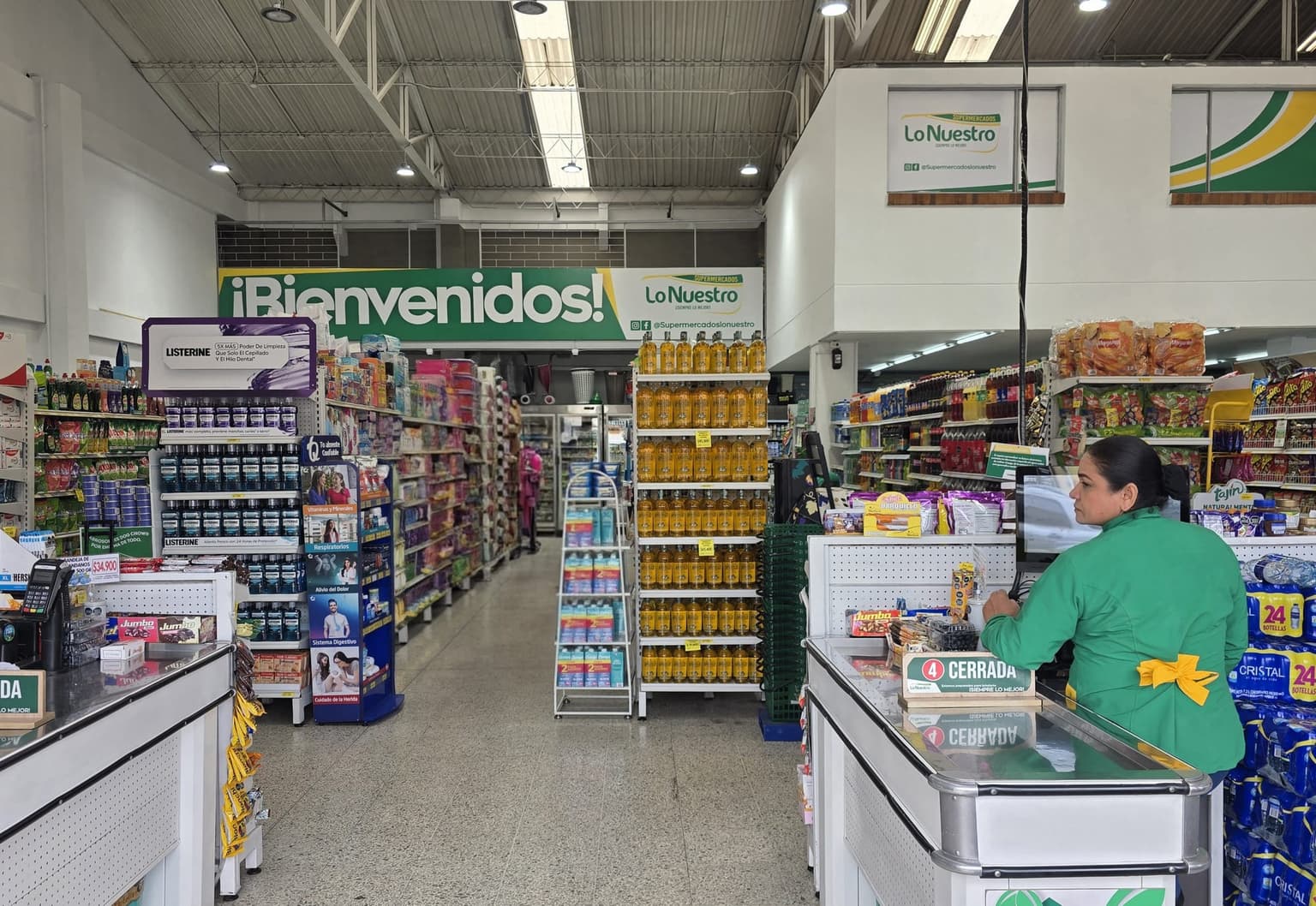 Entrada de una sede con el letrero ¡Bienvenidos! y una cajera atendiendo